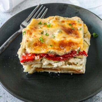 A slice of vegetable lasagna with white sauce on a black plate.