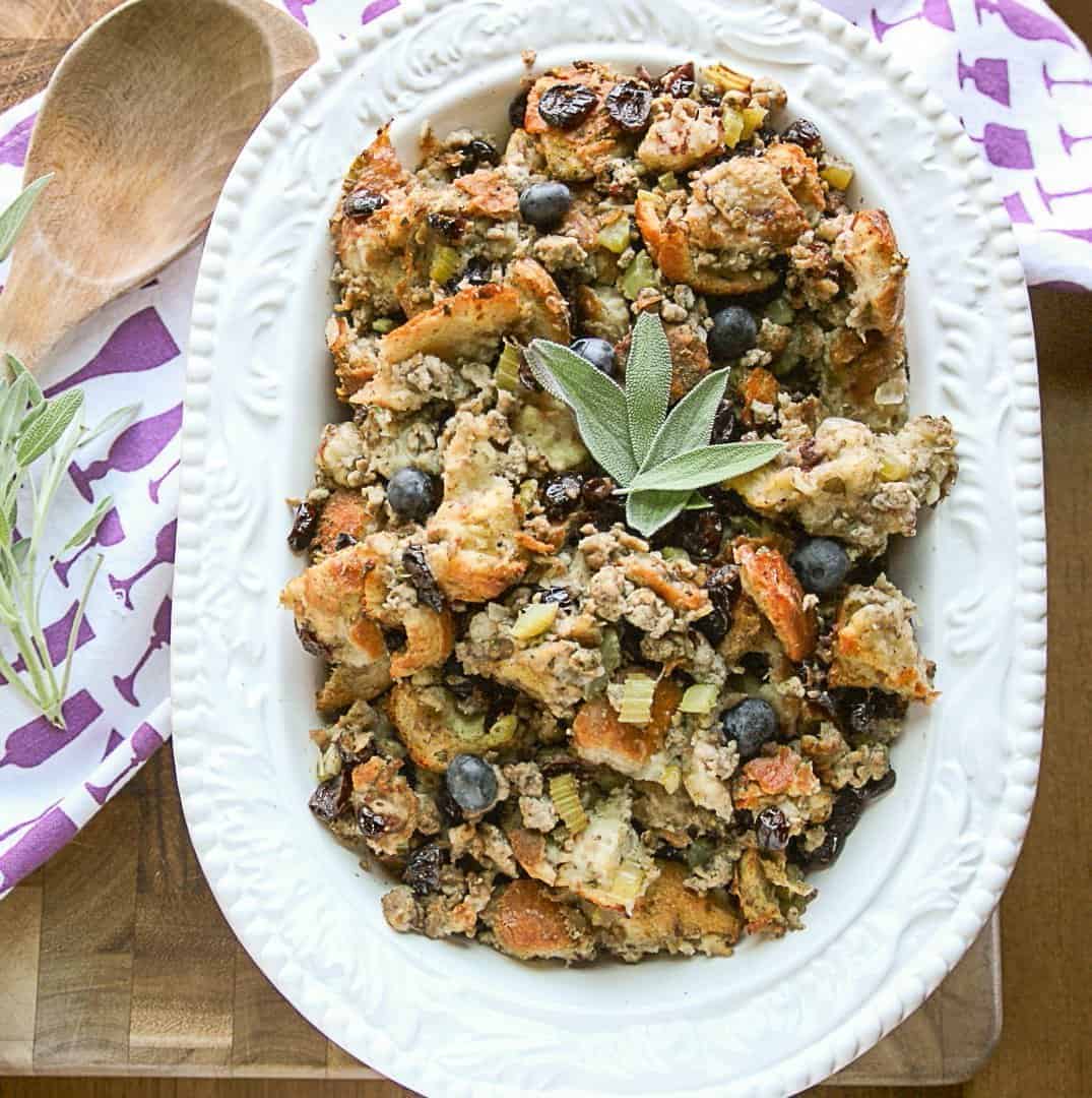 Blueberry sourdough sausage stuffing in a white embossed serving dish with a wooden spoon