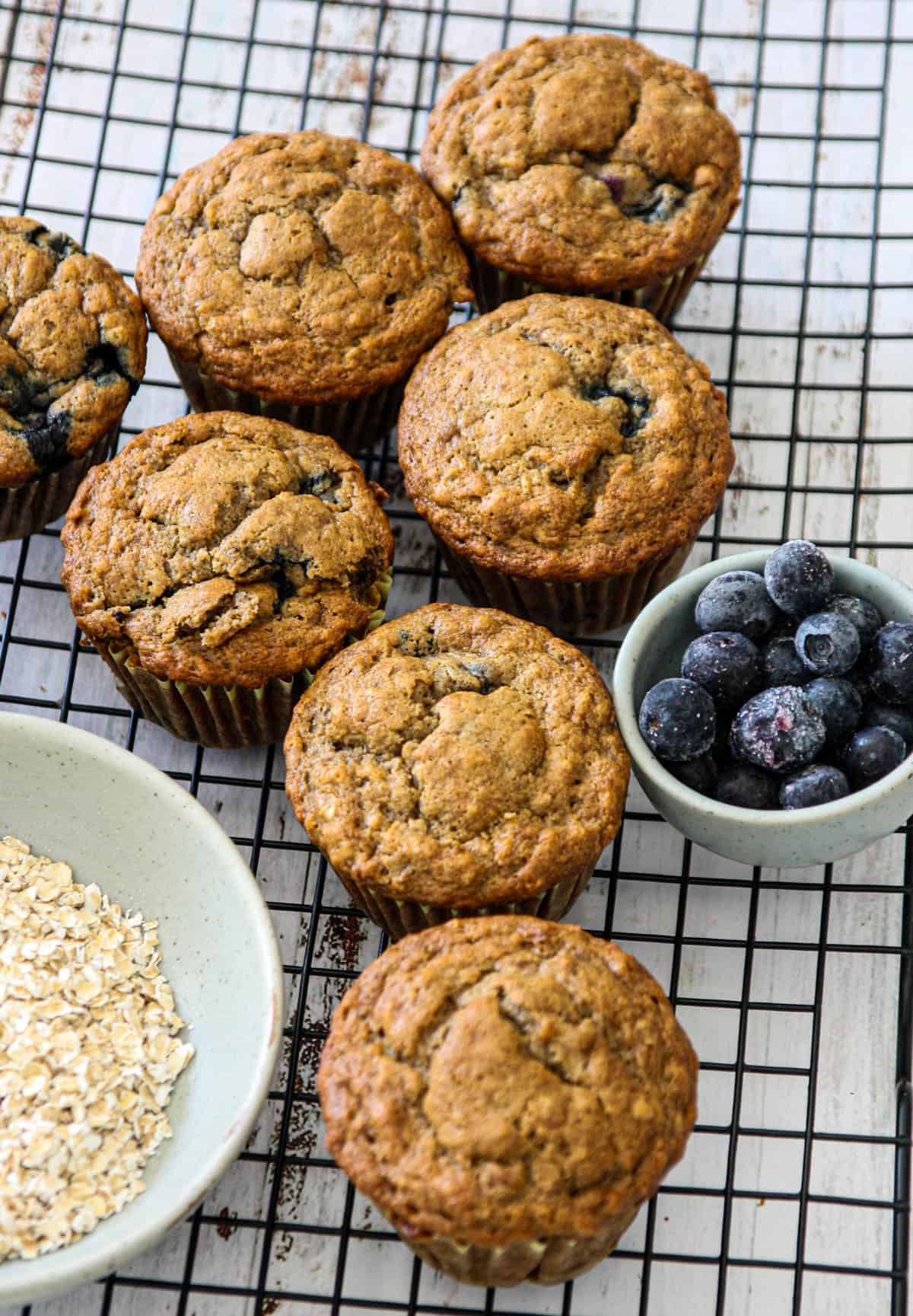 Banana blueberry oatmeal muffins on a wire rack with small dishes of blueberries and oats.