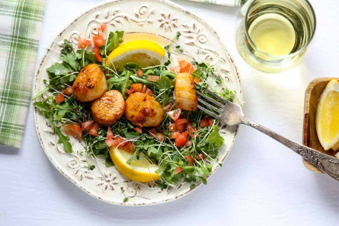 Four large seared scallops on a bed of microgreens on a white embossed plate with two lemon wedges