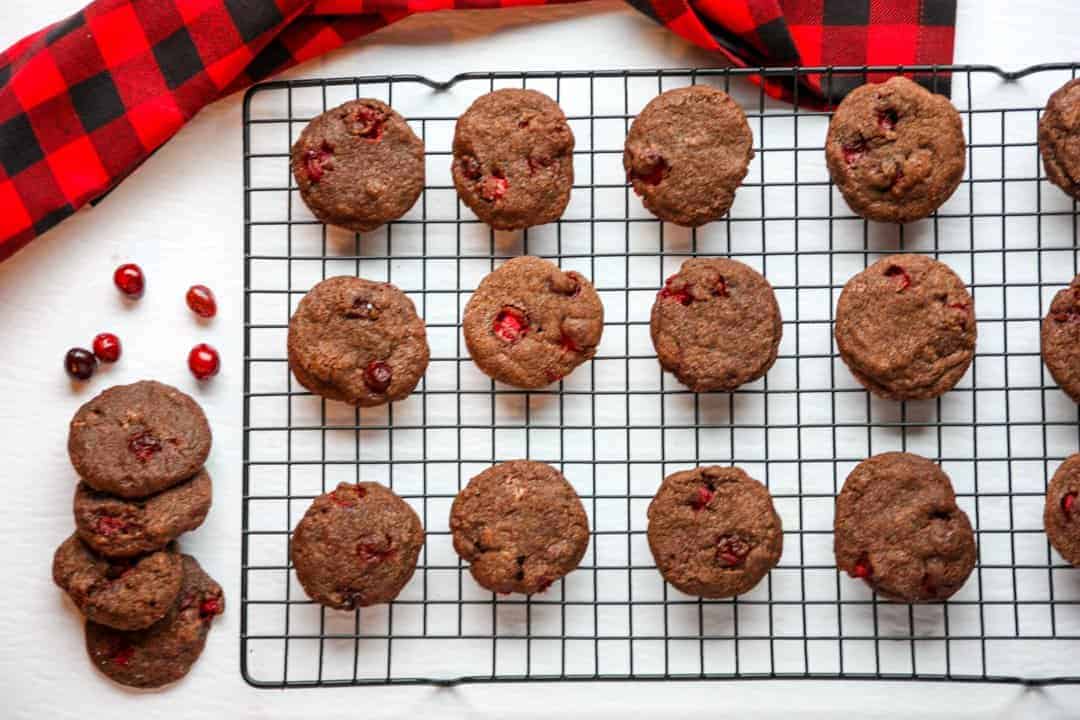 Double chocolate cranberry cookies cooling on a wire baking rack beside a black & red checkered kitchen towel
