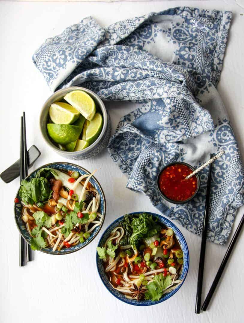 top shot of pho in two bowls next to black chopsticks, a bowl of lemon wedges, and a blue kitchen towel