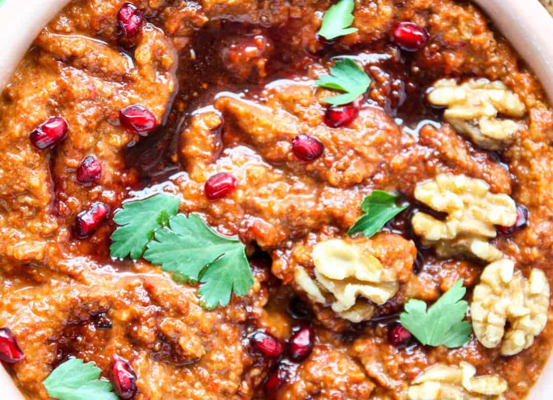 close up of muhammara, a roasted red pepper dip, topped with walnuts, pomegranate arils, and drizzled with pomegranate molasses