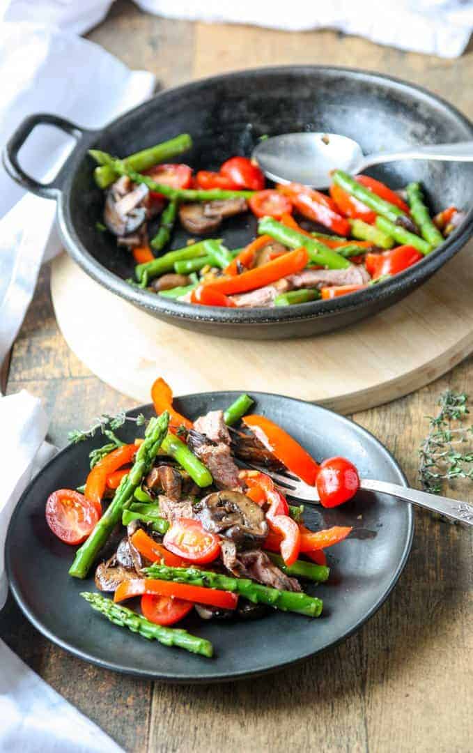 Cast ron pan with steak & asparagus stir fry and one serving of stir fry on a black plate