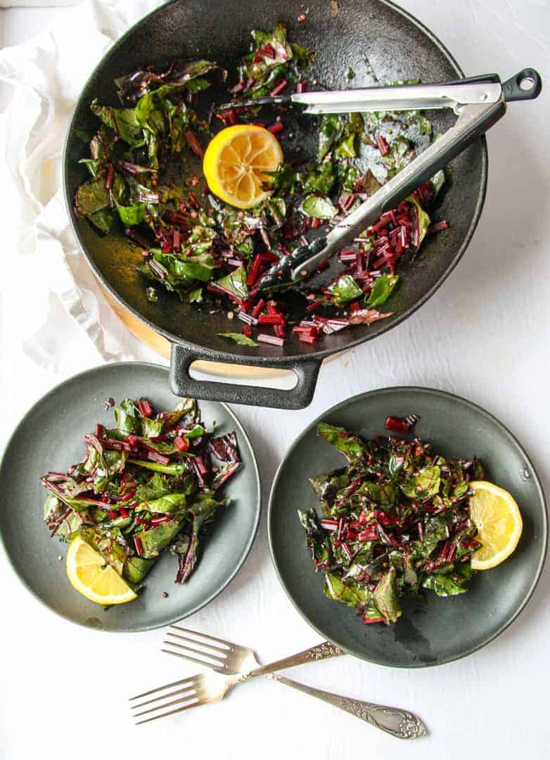 Sautéed Beet Greens being served from a pan onto two plates with tongs.