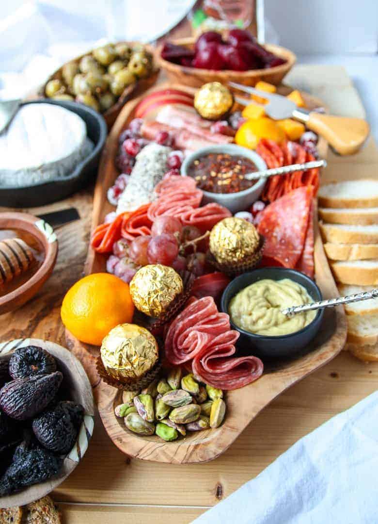 A wooden board full of sliced meats, cheeses, fruits and nuts, olives and chocolate.