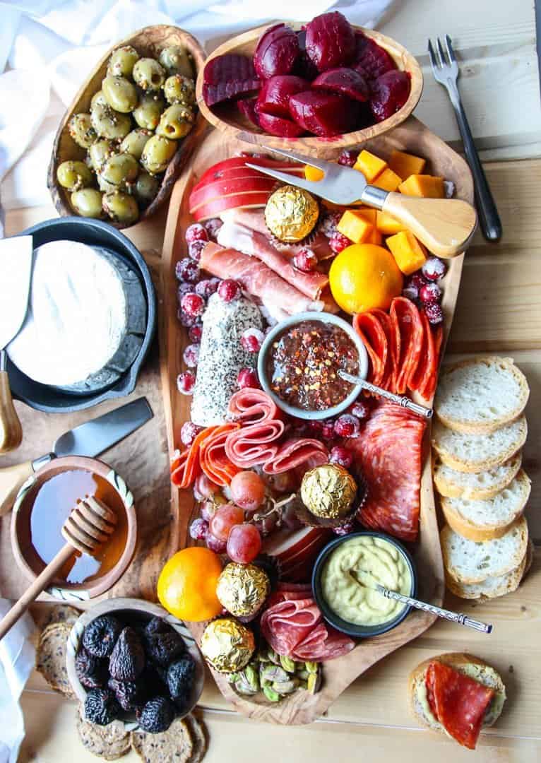 An irregularly shaped wooden charcuterie board filled with meat, cheese, olives, nuts and fruit, surrounded by crackers and bread.