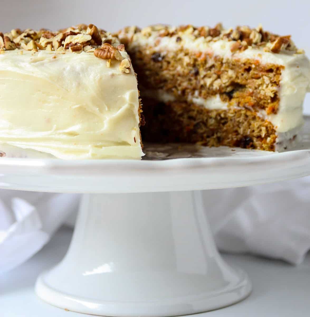 A carrot cake with cream cheese frosting on a cake stand with a slice removed.