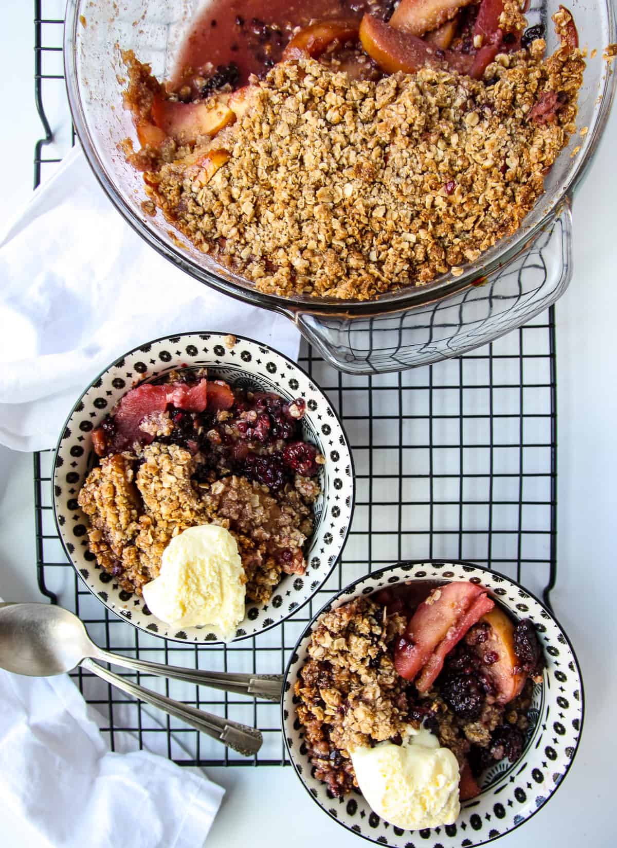 Blackberry and apple crisp in a baking pan and two servings in dishes on a wire rack.