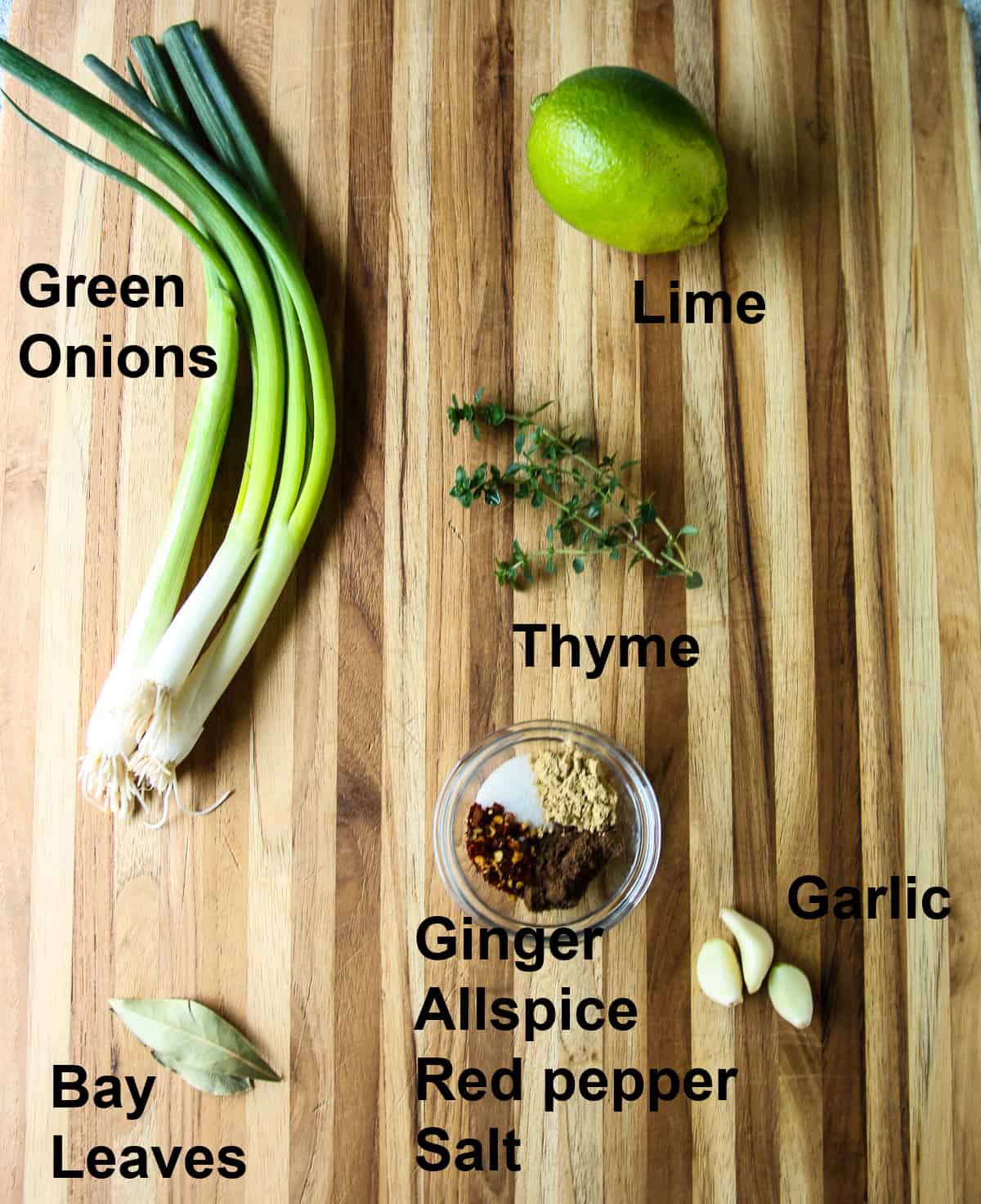 Scallions, garlic cloves, thyme sprigs, bay leaves and a small glass dish of spices on a wooden board.