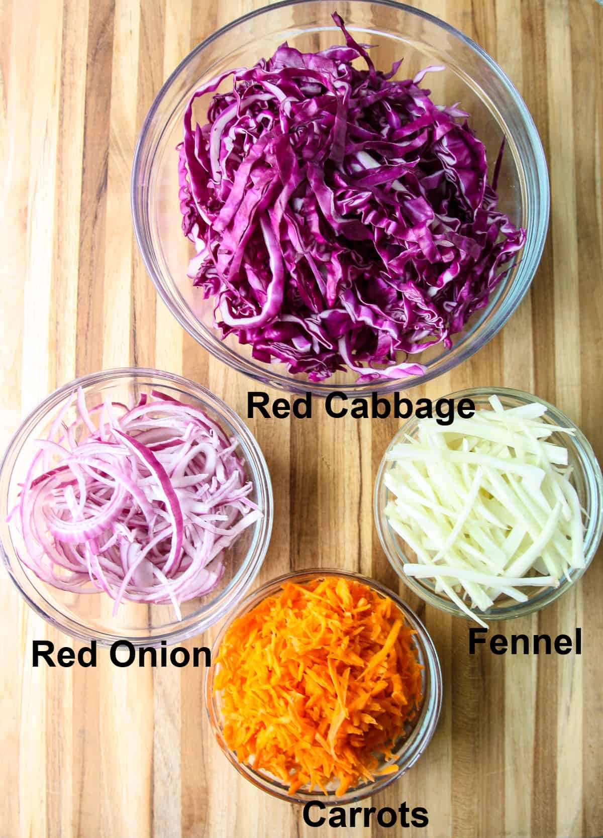 The ingredients to make red cabbage slaw in glass bowls on a wooden board.