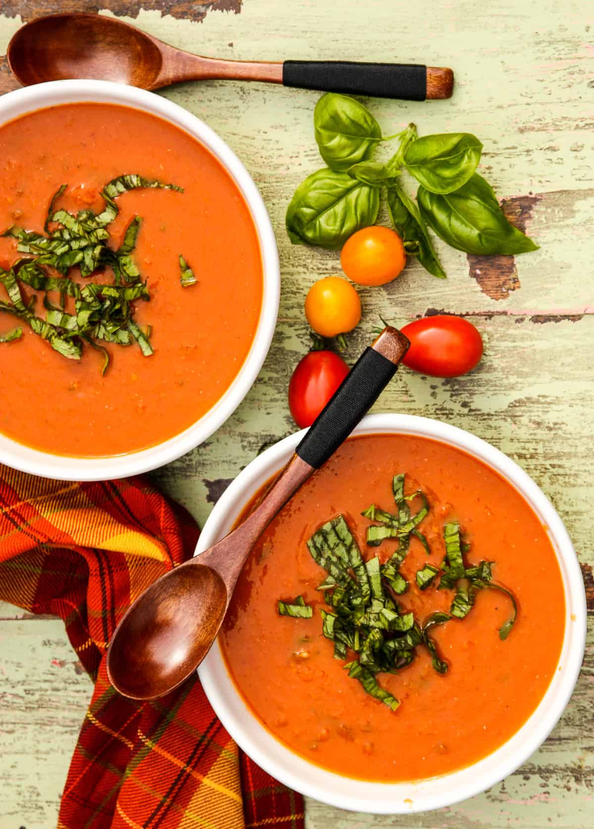Two white bowls filled with roasted tomato basil soup on a table scattered with tomatoes.