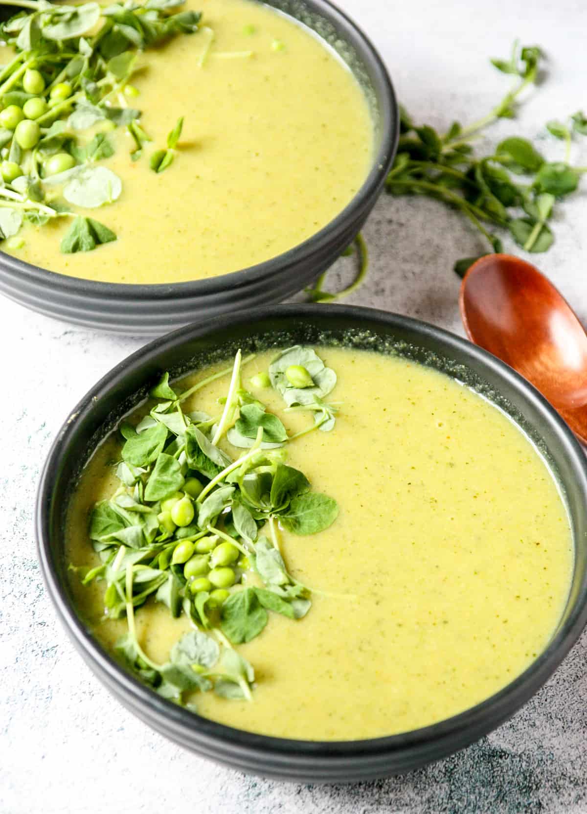Two bowls of broccoli and pea soup, garnished with pea shoots and fresh peas.